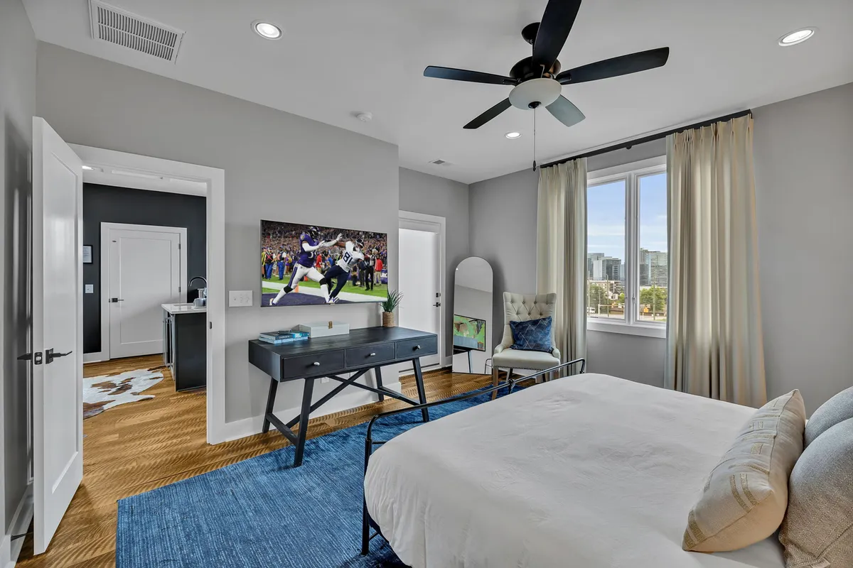 A modern bedroom featuring a king bed with decorative pillows, a large TV displaying a sports scene, a dedicated workspace with a desk and chair, and floor-to-ceiling windows allowing ample natural light. The space is designed for both relaxation and productivity, catering to leisure and business travelers alike.