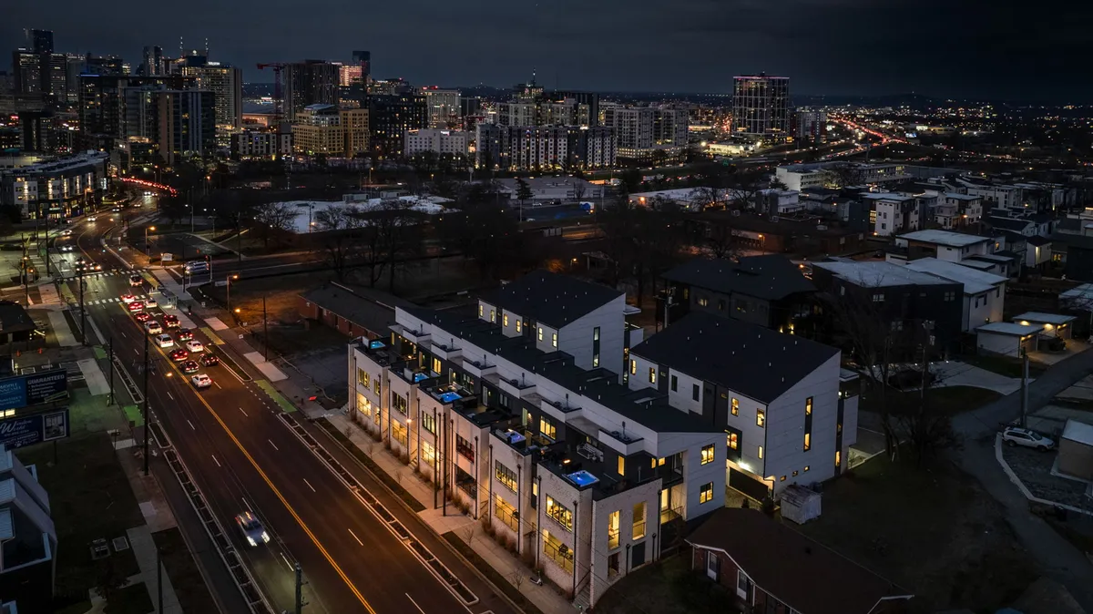 A dynamic exterior view of modern townhouses with the city skyline glowing in the background, lit by streetlights. The setting hints at lively evenings, walkable streets, and the buzz of a sought-after neighborhood.