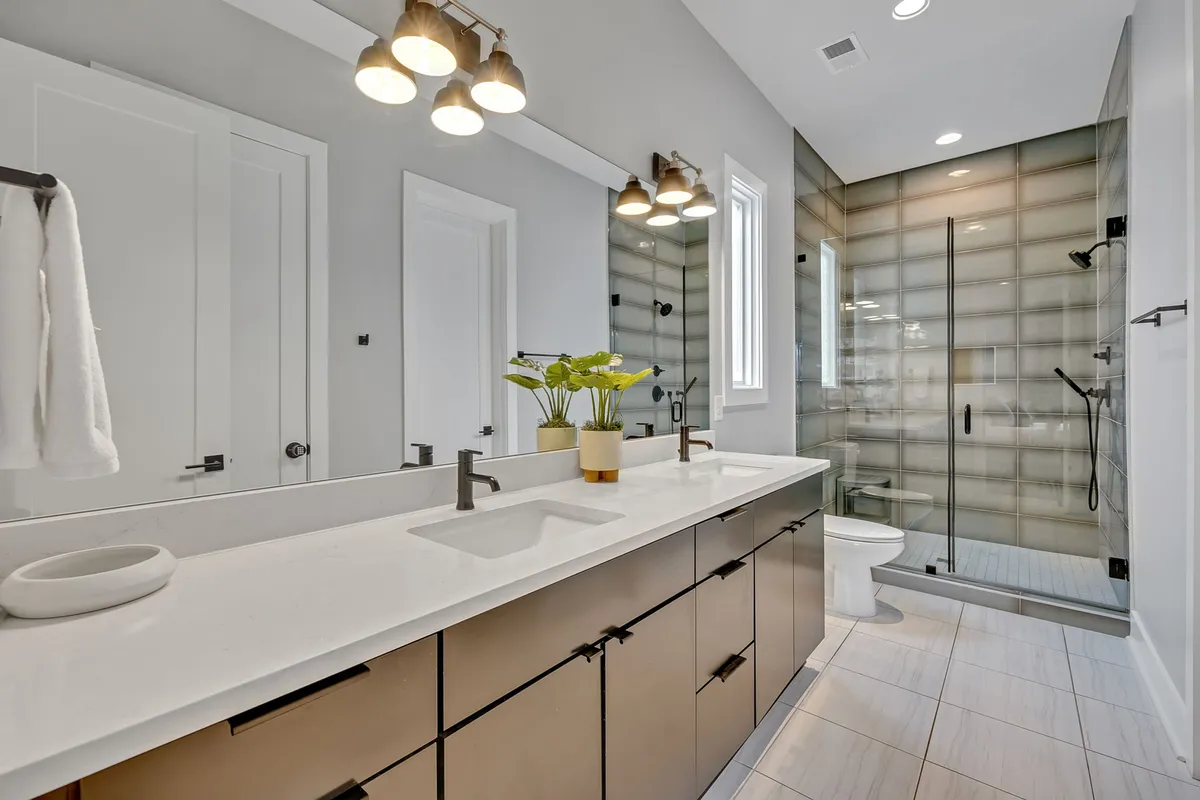 A spa-inspired bathroom with a double sink vanity, glass shower enclosure, decorative plant, and modern lighting fixtures. The setup evokes a sense of calm and luxury, ideal for rejuvenating routines.
