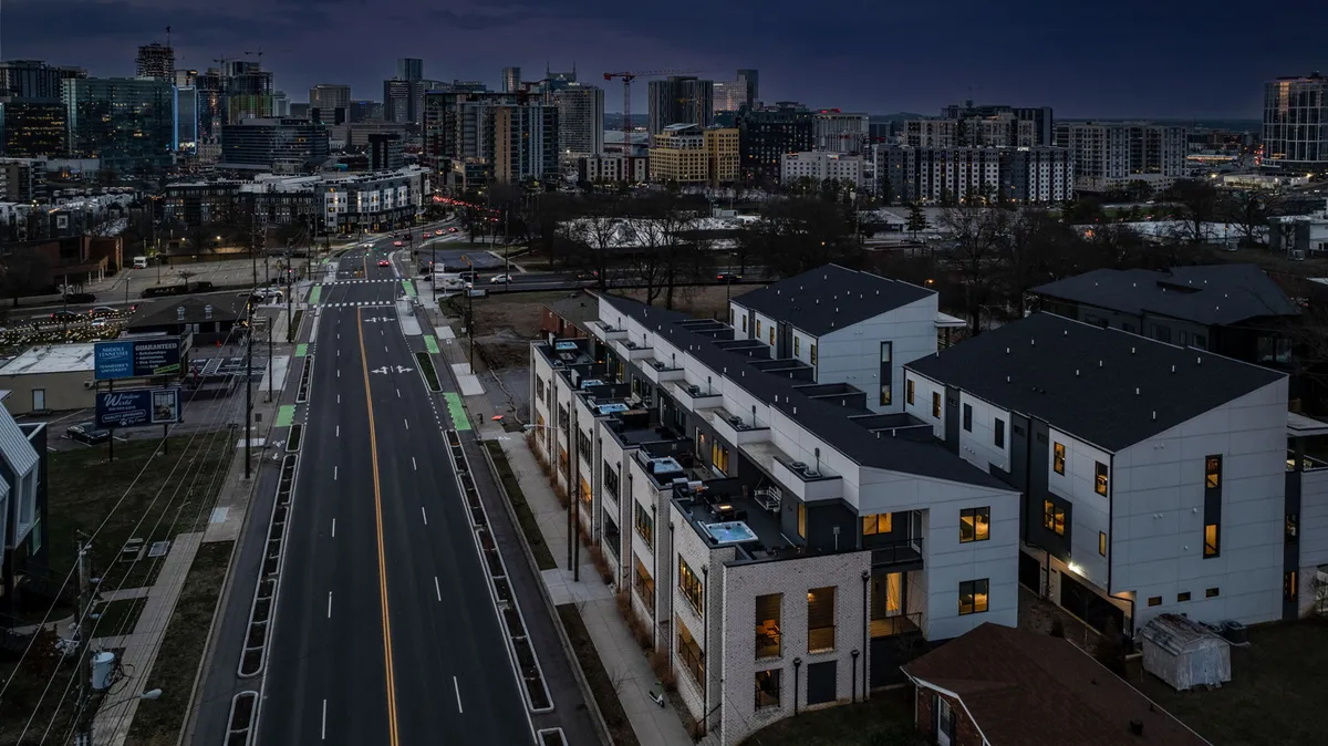 The image depicts contemporary townhouses lining a vibrant city street, with a view of the city skyline in the background and bike lanes in the foreground. It suggests accessibility, modern living, and a lively urban atmosphere perfect for those visiting for work and leisure.