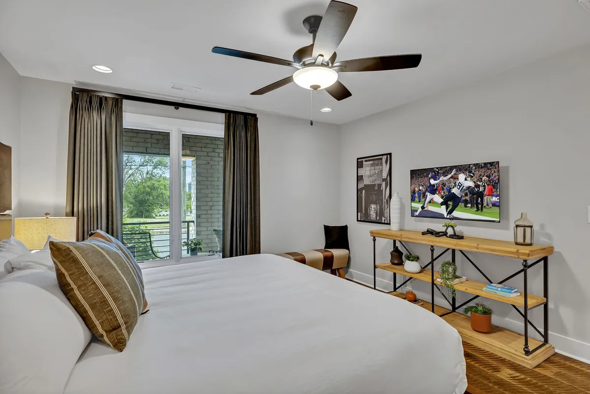 A cozy, contemporary bedroom with a comfortable bed adorned with decorative pillows, a TV mounted on the wall, a wooden shelving unit, and large windows with curtains. The ambiance is modern and inviting, perfect for unwinding after a day in the city.