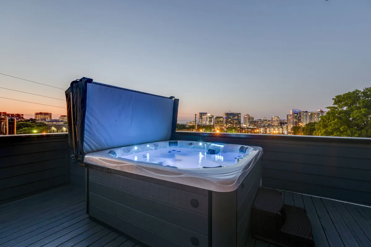 This image captures a rooftop hot tub set against a backdrop of the Nashville skyline at twilight. The city lights shimmer in the distance as guests relax in the bubbling waters, with the open sky above creating a serene and luxurious mood. The deck features comfortable lounge seating, inviting both relaxation and social gatherings.