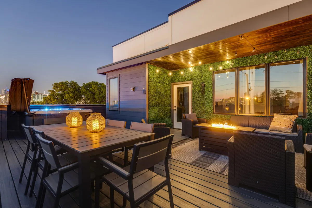 The image showcases a beautifully styled rooftop terrace in downtown Nashville, featuring a large dining table adorned with lanterns, a cozy outdoor seating area centered around a fire pit, and a lush green wall. The space invites gatherings, with city views providing a dramatic backdrop and soft lighting creating a warm, inviting ambiance perfect for evening relaxation or lively dinners with friends and family.